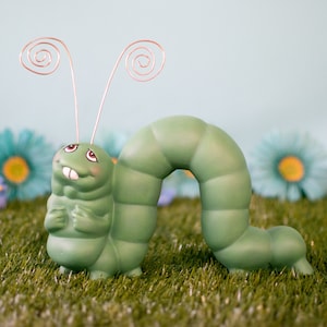 Könnte beinhalten: Eine grüne Keramik-Raupenfigur mit einem skurrilen Ausdruck und Drahtantennen. Die Raupe ist in einer gekrümmten Position platziert, als würde sie gleich kriechen. Die Figur steht auf einem Bett aus grünem Gras.