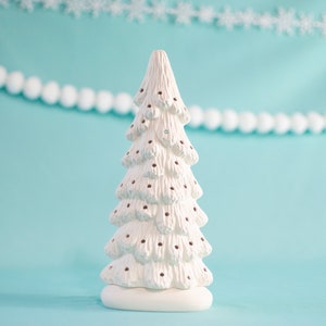 May include: A white ceramic Christmas tree with a textured surface and dark brown dots. The tree is on a white base, with a light blue background and a string of white pom-poms and snowflakes.