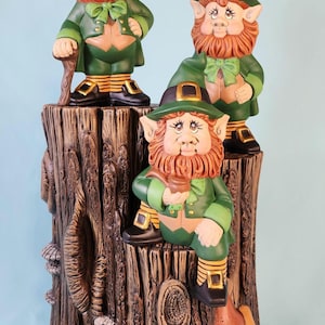 May include: Three ceramic leprechaun figurines sitting on a tree stump with mushrooms. The leprechauns are wearing green hats, green jackets, and have red beards.