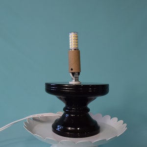 Könnte beinhalten: Ein schwarzer Lampensockel im Sockelstil mit einer weißen Platte mit gewelltem Rand. Eine zylindrische LED-Glühbirne ist in eine Papphülse eingesetzt und auf dem Sockel montiert. Die Lampe steht vor einem türkisfarbenen Hintergrund.