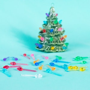 May include: A small ceramic Christmas tree with colorful lights, surrounded by a scattering of replacement light bulbs in various colors, including red, blue, green, yellow, and purple.