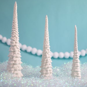 May include: Three white ceramic Christmas trees of varying sizes on a bed of iridescent glitter. The trees are all in a pointed shape with a layered design.