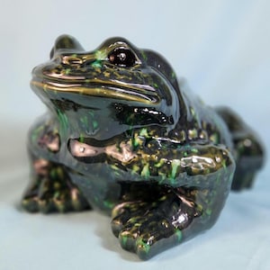 May include: A ceramic frog figurine with a green and black glaze. The frog is sitting with its legs tucked underneath its body and its head tilted up.
