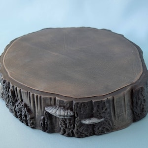 May include: A dark brown wooden tree stump slice with a smooth, flat top surface. The edges of the stump are rough and textured, with small, white mushroom-like details.
