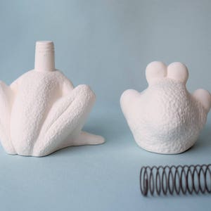 Pu&ograve; includere: Due statuine di rana in ceramica bianca, una con una molla attaccata alla base, e una molla separata.