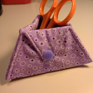 May include: A purple fabric scissor holder with a white floral pattern and a blue button closure. The holder is filled with a pair of orange scissors.