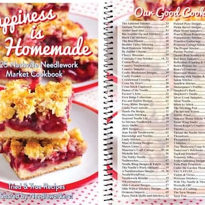 May include: A cookbook titled "Happiness is Homemade" with a 2025 Nashville Needlework Market Cookbook subtitle. The cover features a close-up of a cherry crumble dessert on a white plate with a red rim. The book is open to a page listing "Our Good Cooks" with a list of vendors and their corresponding page numbers.