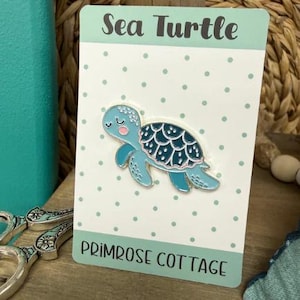 May include: A sea turtle enamel pin with a light blue body and a darker blue shell. The pin is on a white card with a polka dot pattern and the words "Sea Turtle" and "Primrose Cottage".