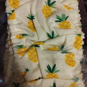 May include: A white fabric diaper cover with a pineapple print. The cover is made of a soft, stretchy material and has a gathered elastic waistband.