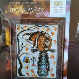 May include: A cross-stitch pattern featuring a beaver with a blue and black tail, surrounded by autumn leaves and a small tent. The pattern is titled "The Beaver" and is part of the "A Year in the Woods" series by Cottage Garden Samplers.
