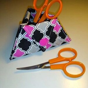 May include: A pair of orange-handled scissors next to a patterned fabric scissor holder. The holder features a black, pink, and gray geometric design. One pair of scissors is inside the holder, and another is lying on the surface.