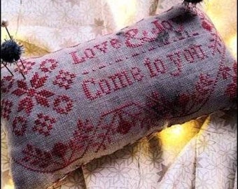 May include: A cross-stitch pillow with a beige background and red stitching. The pillow reads "Love & Joy - Come to you". The pillow is on a white fabric with a snowflake pattern.