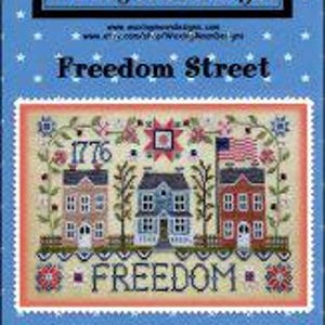 May include: A cross-stitch pattern titled Freedom Street by Waxing Moon Designs. The design features three houses, the year 1776, an American flag, and the word FREEDOM. The background is light blue with white stars, and the pattern is labeled #287.