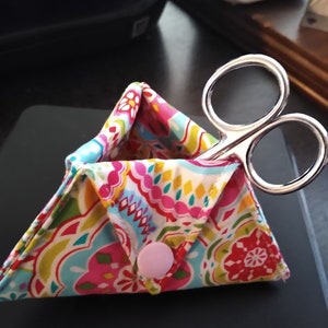 May include: A small, triangular fabric pouch with a colorful paisley pattern. The pouch has a pink button closure and is holding a pair of silver scissors.