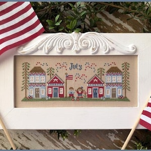 May include: A framed cross-stitch featuring two houses, a flag, and the word "July." The houses are red and blue with white trim. Two American flags are on either side of the frame. The frame is white with a decorative top.