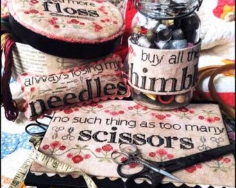May include: A collection of cross-stitch supplies, including a jar labeled "buy all the thimbles", a needle case, and a round container with "need more floss" text. Scissors and measuring tape are also visible.