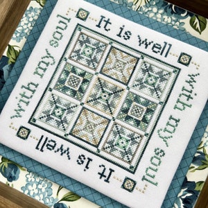May include: A cross-stitch piece with the words "It is well with my soul" in green. The design features a square pattern in shades of green, beige, and white. The piece is displayed on a blue patterned fabric.