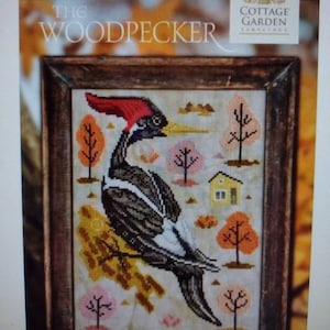 May include: A cross-stitch pattern featuring a black and white woodpecker with a red head perched on a branch. The background is a colorful autumn scene with trees and a small house.