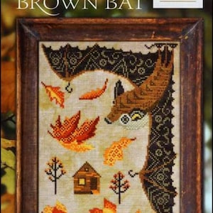 May include: A framed cross-stitch pattern featuring a brown bat with a cabin and autumn leaves. The title of the pattern is "The Little Brown Bat" and is part of the "A Year in the Woods" series, number 10. The pattern is from Cottage Garden Samplings.