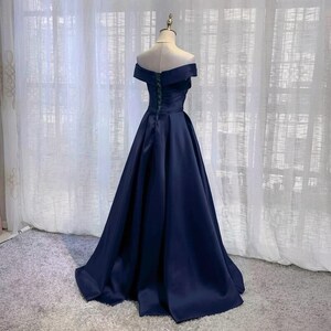 May include: A navy blue off-the-shoulder gown with a full skirt. The dress features a fitted bodice and a flowing skirt. The back of the dress has a green lace-up detail. The dress is displayed on a mannequin in front of a sheer white curtain.
