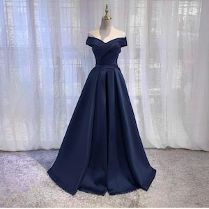 May include: A navy blue off-the-shoulder formal gown. The dress features a fitted bodice with a sweetheart neckline and a flowing A-line skirt. The fabric has a subtle sheen, and the dress is displayed on a mannequin against a soft, neutral background.