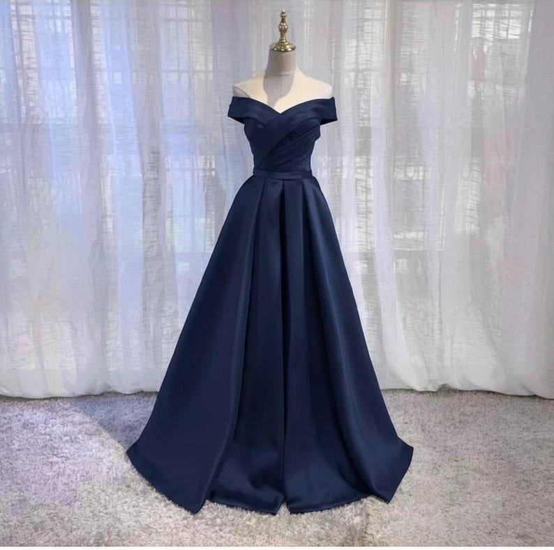 May include: A navy blue off-the-shoulder formal gown. The dress features a fitted bodice with a sweetheart neckline and a flowing A-line skirt. The fabric has a subtle sheen, and the dress is displayed on a mannequin against a soft, neutral background.