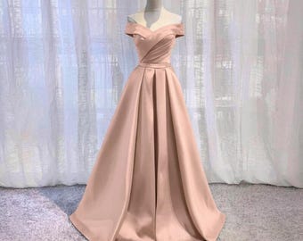 May include: A blush-colored, off-the-shoulder formal gown. The dress features a fitted bodice with a sweetheart neckline and a pleated, full A-line skirt. The fabric has a satin sheen, and the dress is displayed on a mannequin.