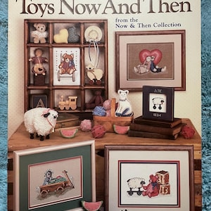 May include: A craft book cover titled "Toys Now And Then" featuring cross-stitch designs. The image showcases framed cross-stitch samplers, a wooden shelf with miniature toys, and decorative elements like watermelon slices and a sheep figurine.