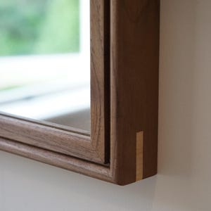 May include: A rectangular wall mirror featuring a dark brown wooden frame with a visible grain pattern. The corner of the frame displays a contrasting light-coloured wood inlay detail, showcasing a clean mitered joint construction.