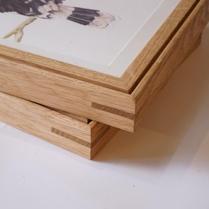 May include: Close-up of two wooden picture frames, one stacked on top of the other. The frames are made of light-coloured wood with visible grain and intricate corner joinery. The top frame holds a watercolour painting of a bird.