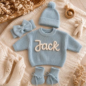 May include: A light blue knitted baby set consisting of a long-sleeve sweater with the name Jack embroidered in white, a matching beanie with a pom-pom, a bow headband, and a pair of tie-up booties, all displayed on a textured cream fabric.