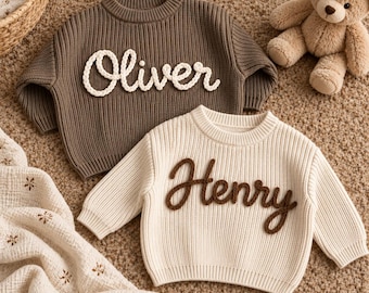 May include: Two long-sleeved knit sweaters. One is dark brown with the name Oliver in white yarn lettering. The other is cream-colored with the name Henry in dark brown yarn lettering. Both feature a ribbed crew neck and cuffs.