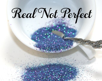 May include: A white ceramic bowl filled with blue and purple glitter, with a small spoon resting inside. The words "Real Not Perfect" are written in black script across the top of the bowl. Glitter is also scattered on the white surface.