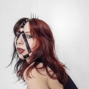 Op de afbeelding: Een zwart lederen hoofddeksel met een reeks zilverkleurige metalen spikes aan de bovenkant en een geometrisch bandontwerp dat het gezicht omlijst. Het accessoire bevat cirkelvormige metalen ringen bij het voorhoofd en de kin, verbonden door verstelbare banden.