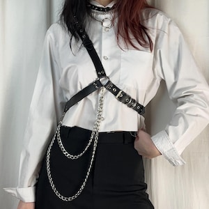 May include: Black leather body harness with silver-tone chain accents. The harness features adjustable straps and a buckle closure. A silver chain dangles from the center of the harness. A matching black choker necklace with a silver ring is also visible.