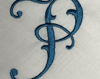 Könnte beinhalten: Eine weiße Stoffoberfläche mit einem großen, dekorativen Buchstaben P, der in tiefem blaugrünem Garn gestickt ist. Die Stickerei zeigt einen fließenden, kalligrafischen Schriftstil mit komplexen Schleifen und Kurven auf dem weißen Textil.