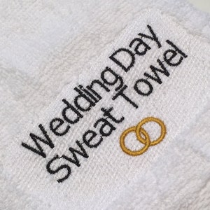 K&ouml;nnte beinhalten: Wei&szlig;es Frottee-Schwei&szlig;handtuch mit dem gestickten Text "Wedding Day Sweat Towel" in Schwarz. Zwei goldene Eheringe sind darunter gestickt. Das Handtuch besteht aus weichem, saugf&auml;higem Material.
