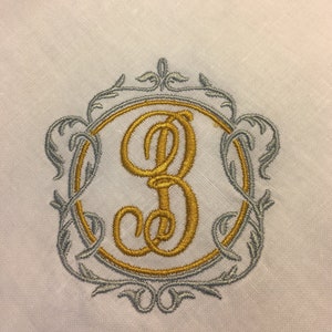 May include: A gold embroidered letter B in a silver ornate frame on a white linen background.