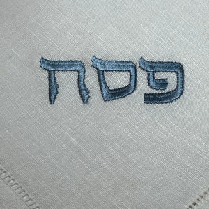 Może przedstawiać: Biała serwetka lniana z haftowanym niebieską nicią hebrajskim słowem "Pesach".