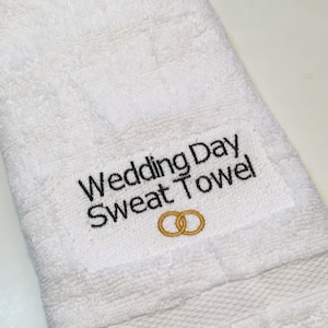 K&ouml;nnte beinhalten: Wei&szlig;es Frotteehandtuch mit dem gestickten Text "Wedding Day Sweat Towel" in Schwarz. Unter dem Text sind zwei goldene Eheringe gestickt. Das Handtuch ist gefaltet und hat eine strukturierte Oberfl&auml;che.