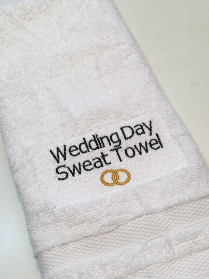 K&ouml;nnte beinhalten: Wei&szlig;es Frotteehandtuch mit dem gestickten Text "Wedding Day Sweat Towel" in Schwarz. Unter dem Text sind zwei goldene Eheringe gestickt. Das Handtuch ist gefaltet und hat eine strukturierte Oberfl&auml;che.