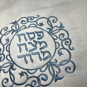 Może przedstawiać: Biały materiał lniany z niebieskim haftem. Haft przedstawia okrąg z hebrajskimi słowami "Pesach Sameach" w ozdobnym kroju pisma, otoczony wzorem kwiatowym.