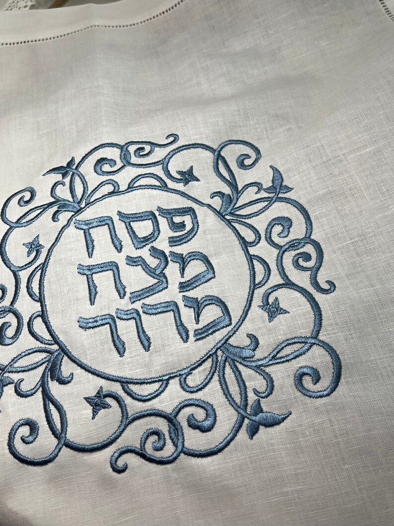 Może przedstawiać: Biały materiał lniany z niebieskim haftem. Haft przedstawia okrąg z hebrajskimi słowami "Pesach Sameach" w ozdobnym kroju pisma, otoczony wzorem kwiatowym.