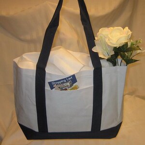 Puede incluir: Bolsa de lona blanca con asas y ribetes azul marino. La bolsa tiene un bolsillo grande en la parte delantera con una peque&ntilde;a tarjeta que dice "Shopping List" en su interior.