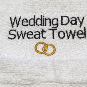 K&ouml;nnte beinhalten: Wei&szlig;es Frottee-Schwei&szlig;handtuch mit dem gestickten Text "Wedding Day Sweat Towel" in Schwarz. Unter dem Text befinden sich zwei goldgestickte Eheringe. Das Handtuch ist f&uuml;r den Hochzeitstag bestimmt.