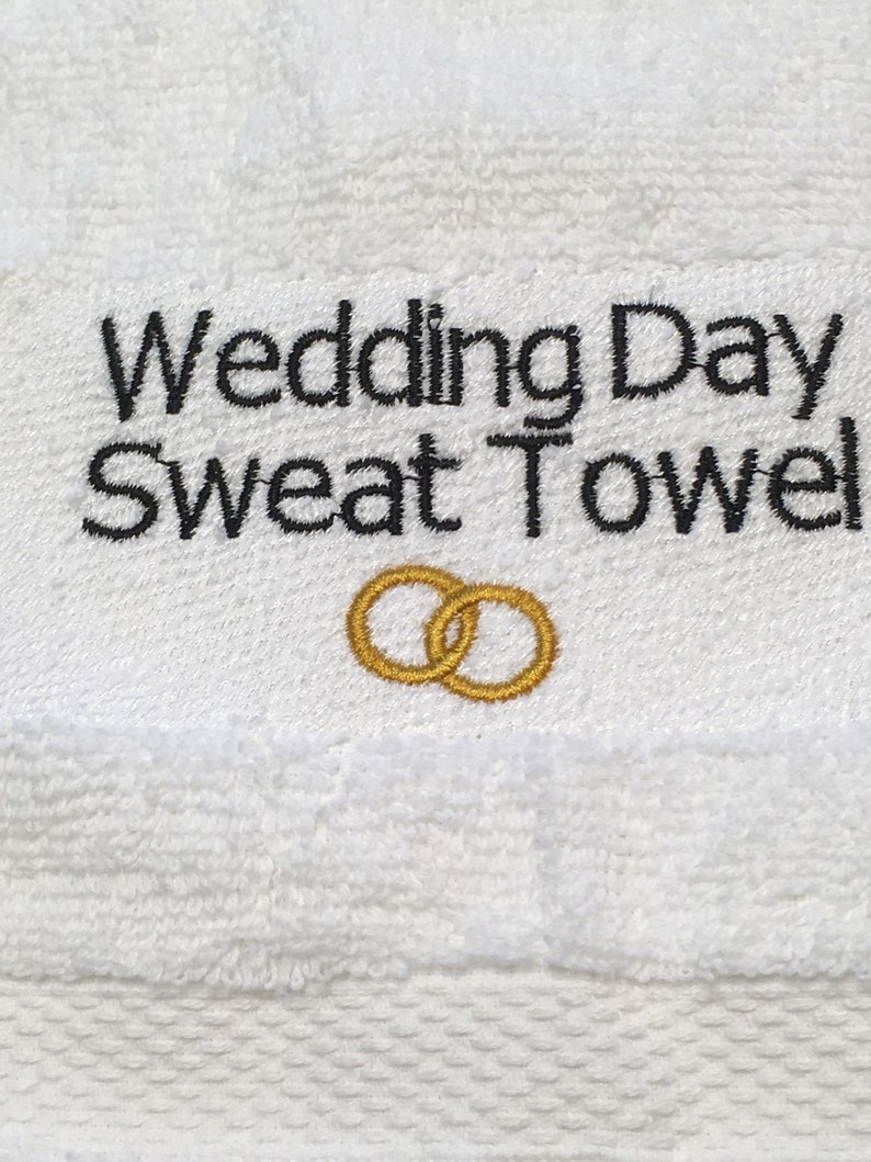 K&ouml;nnte beinhalten: Wei&szlig;es Frottee-Schwei&szlig;handtuch mit dem gestickten Text "Wedding Day Sweat Towel" in Schwarz. Unter dem Text befinden sich zwei goldgestickte Eheringe. Das Handtuch ist f&uuml;r den Hochzeitstag bestimmt.