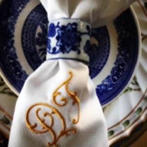 May include: White linen napkin with a gold embroidered initial "D" on a blue and white patterned plate.