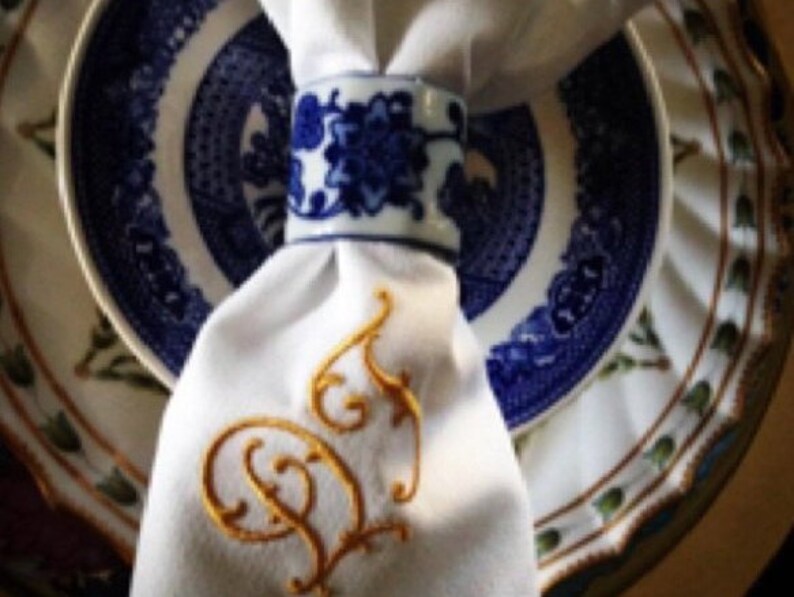 May include: White linen napkin with a gold embroidered initial "D" on a blue and white patterned plate.