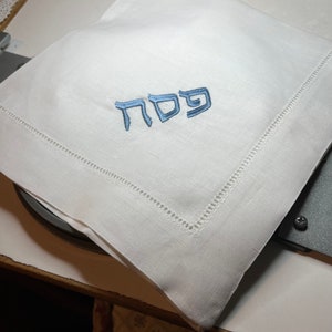 Może przedstawiać: Biała serwetka lniana z niebieskim haftem hebrajskim, kt&oacute;ry brzmi "Pesach"