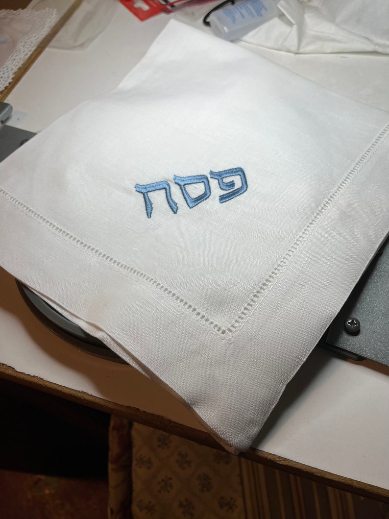 Może przedstawiać: Biała serwetka lniana z niebieskim haftem hebrajskim, kt&oacute;ry brzmi "Pesach"